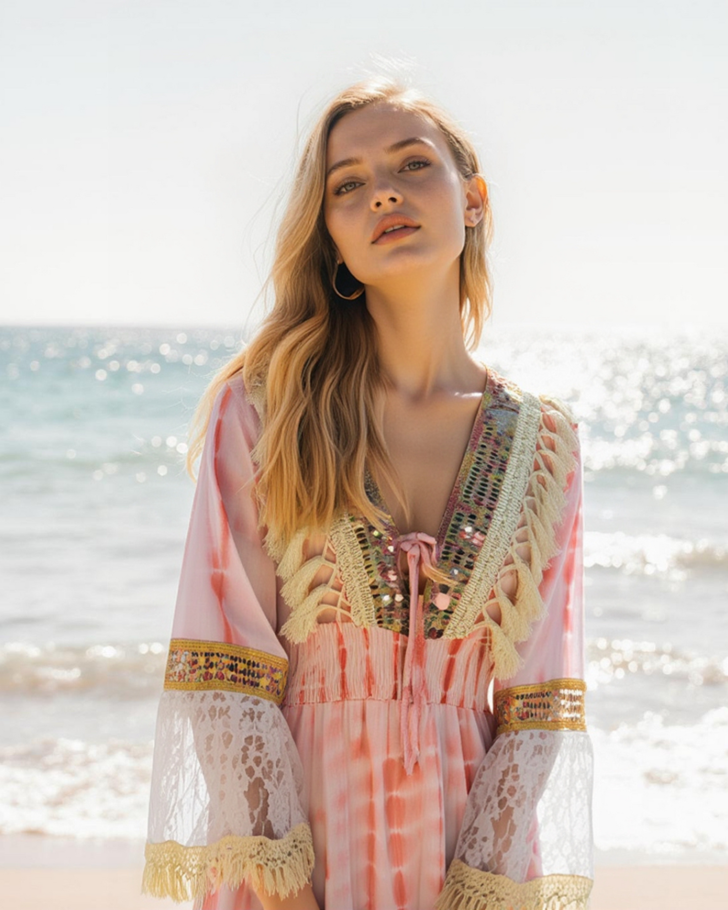 Eine junge Frau mit langen blonden Haaren trägt ein rosafarbenes Boho-Kleid mit Stickereien an einem sonnigen Strand mit glitzerndem Meer im Hintergrund.