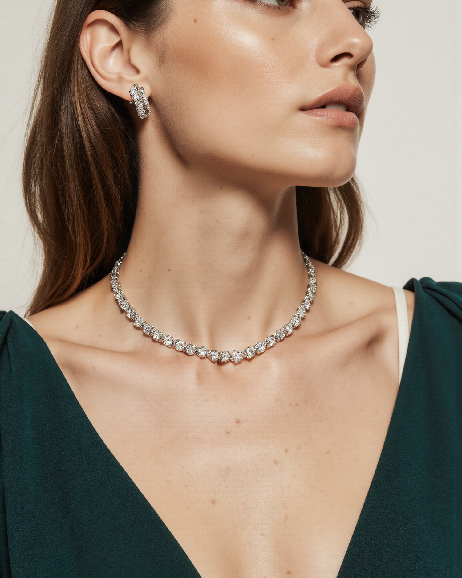 Eine Frau trägt ein elegantes, tief ausgeschnittenes, dunkelgrünes Kleid. Ihr Blick ist nach rechts gerichtet. Sie trägt eine funkelnde Diamant-Halskette, passende Ohrringe und ein dezentes Make-up. 