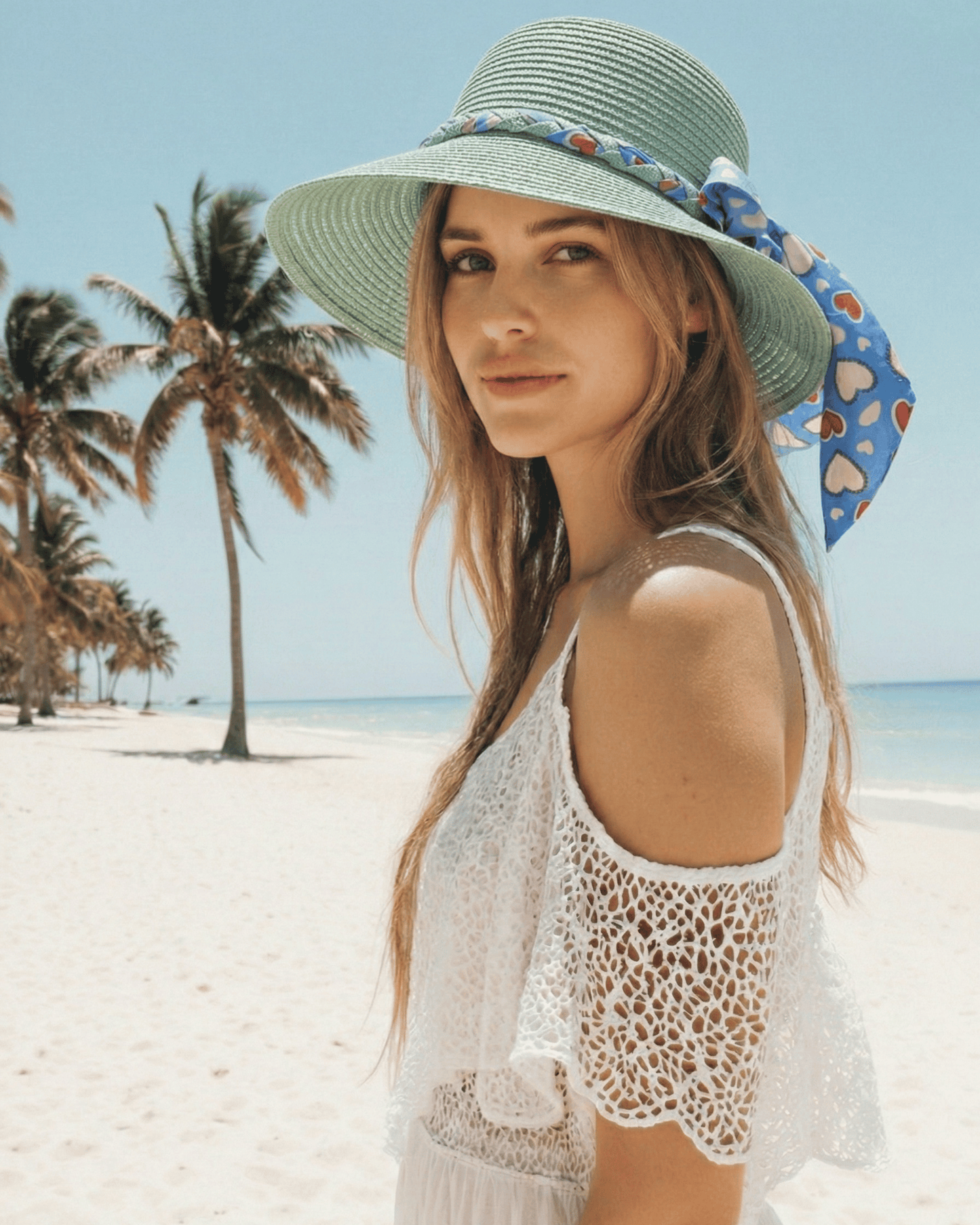 Lächelnde Frau trägt den mintgrünen Strohhut „Amora“ mit Herz-Schleife vor einer tropischen Strandkulisse.