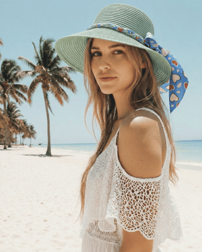 Lächelnde Frau trägt den mintgrünen Strohhut „Amora“ mit Herz-Schleife vor einer tropischen Strandkulisse.