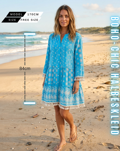 Ganzkörper-Frontansicht einer Frau an einem sonnigen Strand, die ein leuchtend blaues Boho-Mini-Kleid mit aufwendiger Paisley-Stickerei, geometrischen Mustern und Quastenfransen-Details an Ärmeln und Saum trägt. Enthält Grafiken für Free Size, 84 cm Länge und 170 cm Körpergröße, sowie den Text BOHO-CHIC HALBESKLEID.