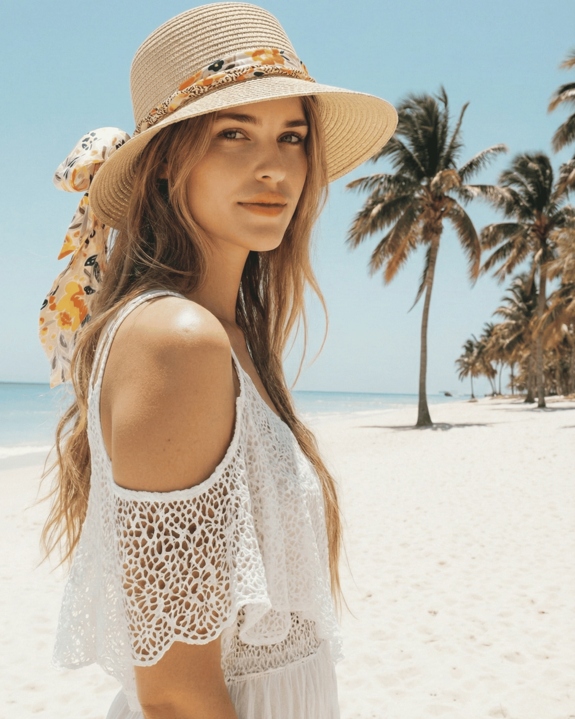 Frau mit blonden Haaren trägt den Strohhut „Liana“ mit floraler Schleife an einem hellen Sandstrand mit Palmen.