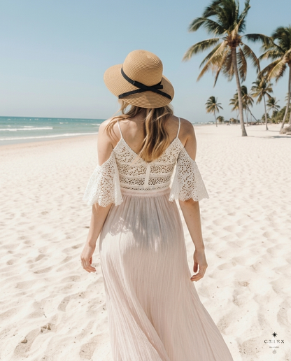 Rückenansicht derselben Frau, die das weiße Boho-Kleid mit den weiten Häkelärmeln und einen Strohhut mit Schleife zeigt, während sie auf den Strand und das Meer blickt.