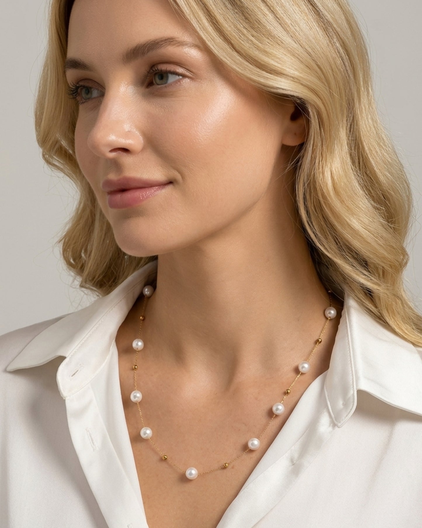 Nahaufnahme einer blonden Frau im weißen Hemd, die die elegante, goldene Halskette mit den schimmernden Perlen trägt.