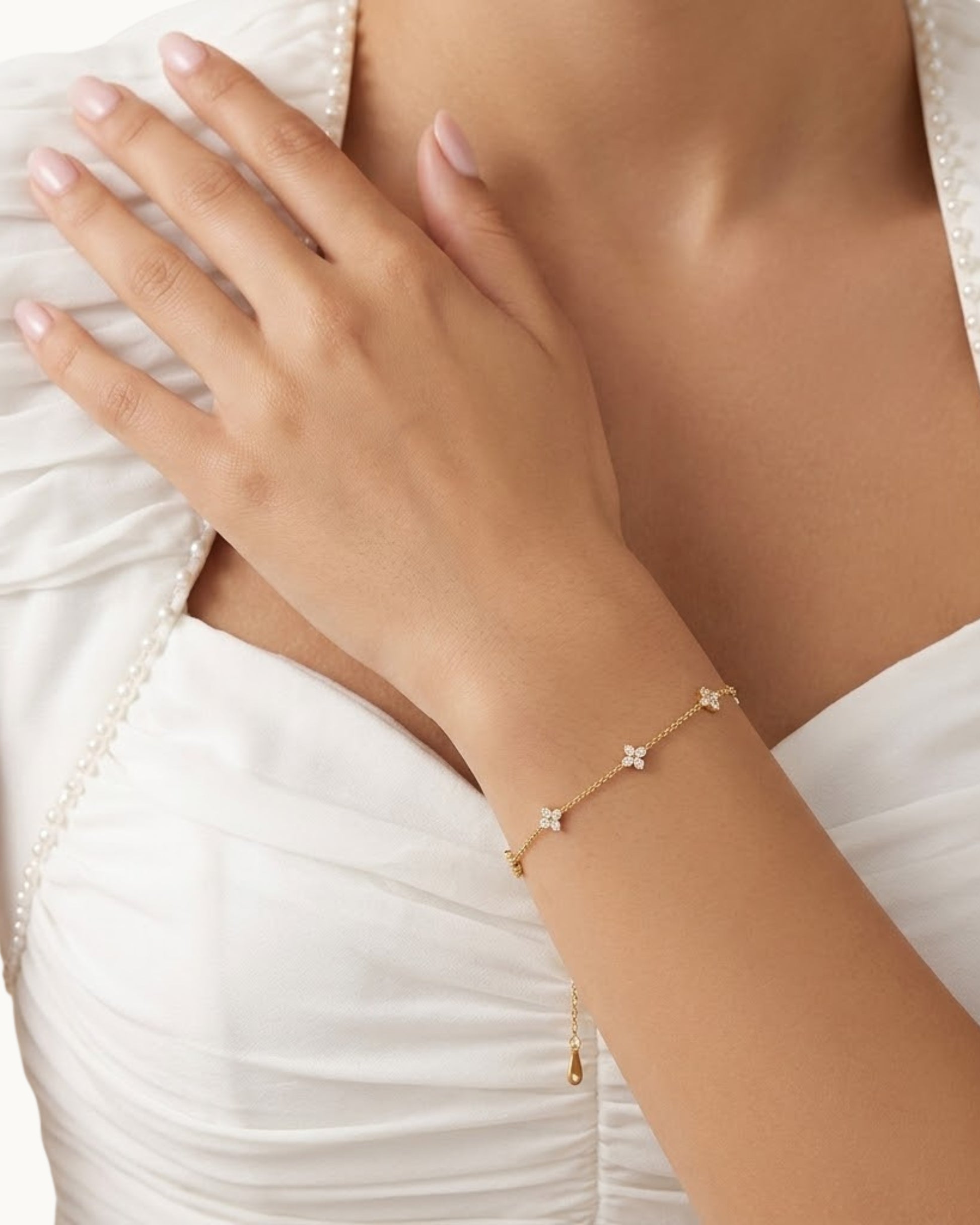 Nahaufnahme eines zarten, goldenen Edelstahl-Armbands mit funkelnden Kleeblatt-Anhängern am Handgelenk einer Frau vor einem weißen Kleid.