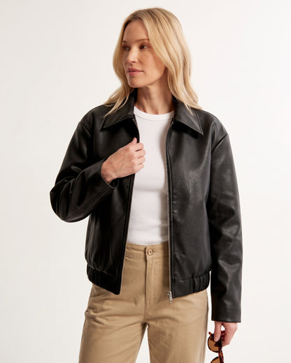 Blondes Model trägt offene schwarze Bikerjacke in genarbter Lederoptik kombiniert mit weißem T-Shirt und beiger Hose